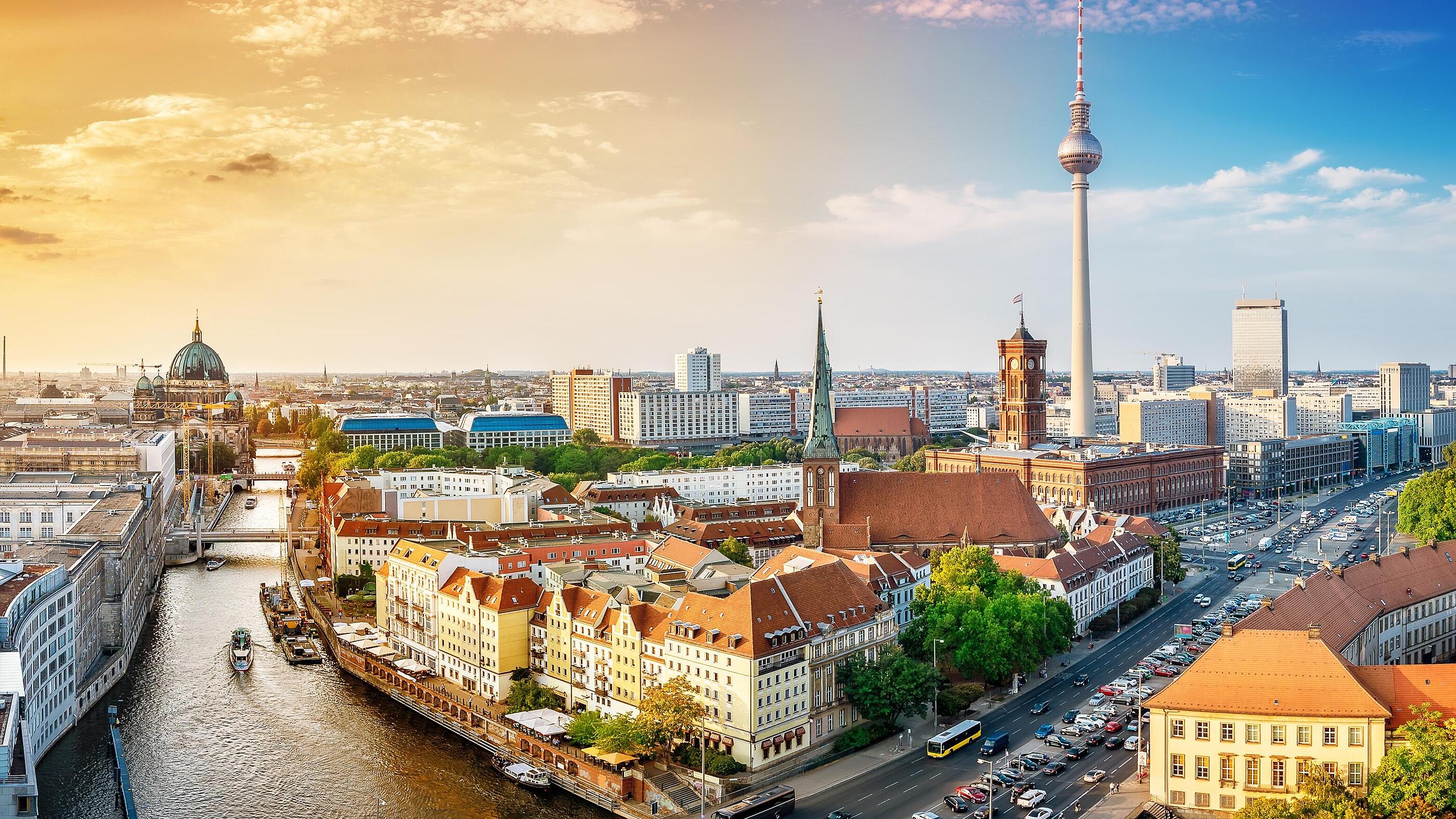 Panoramablick auf Berlin während des Sonnenuntergangs. Die Stadt zeigt eine Mischung aus modernen und historischen Gebäuden, darunter der Berliner Fernsehturm. Ein Fluss fließt durch die Stadt, umgeben von Bäumen und Straßen mit Fahrzeugen.